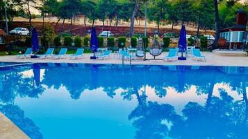 A swimming pool with lounge chairs and umbrellas, surrounded by trees and a building in the background.