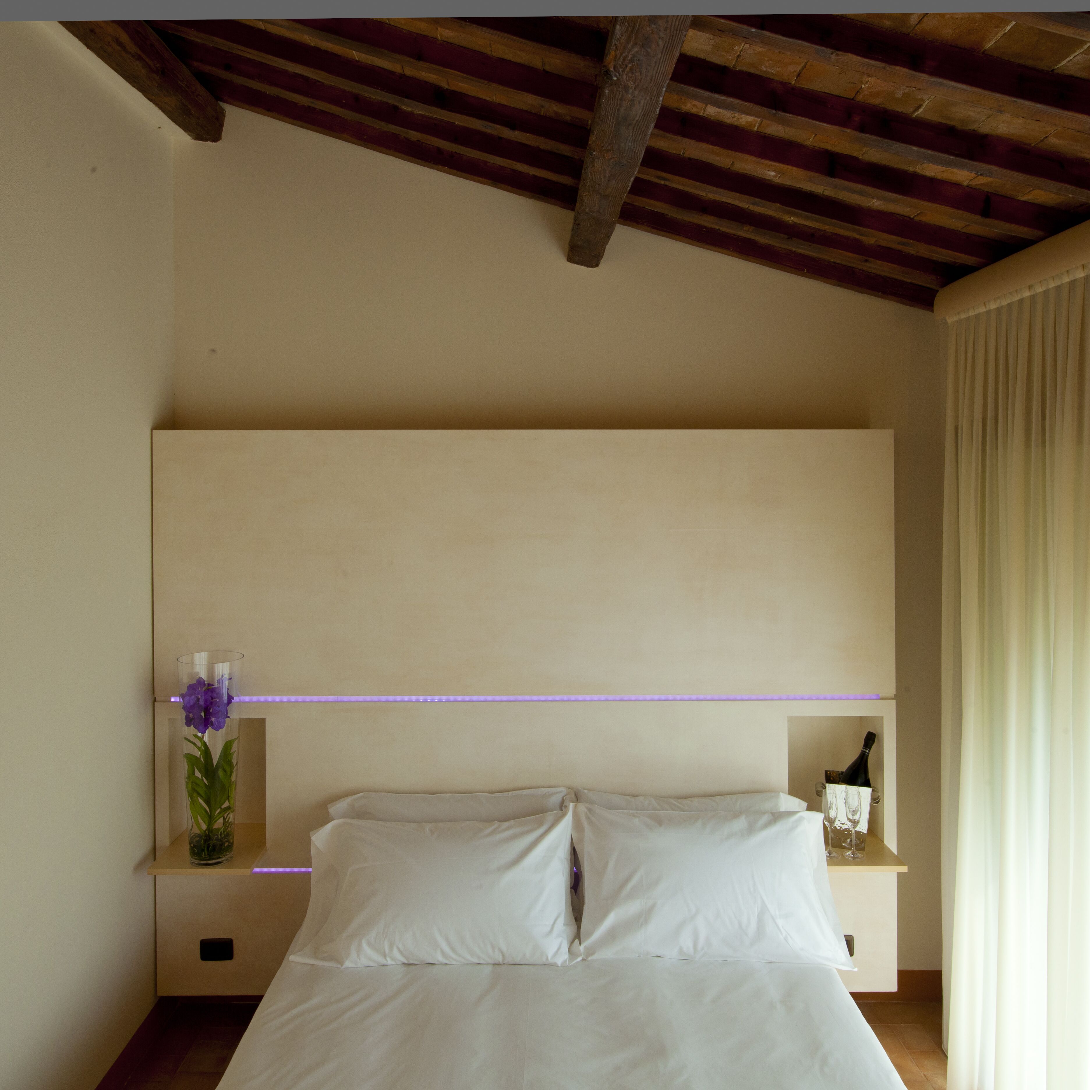 A bedroom with a bed, a nightstand, a wall-mounted shelf with a bottle and glasses, and a purple light strip.