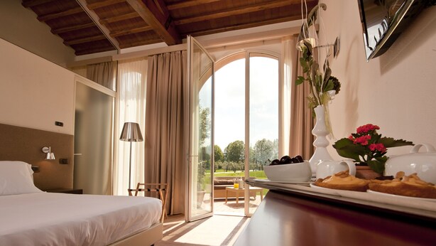 Habitación de hotel con cama, una mesa de comedor con alimentos para el desayuno, una planta en maceta y vistas a un espacio exterior a través de un ventanal.