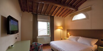 Habitación de hotel con cama, escritorio, televisor, ventana con cortinas, lámpara y techo con vigas a la vista.