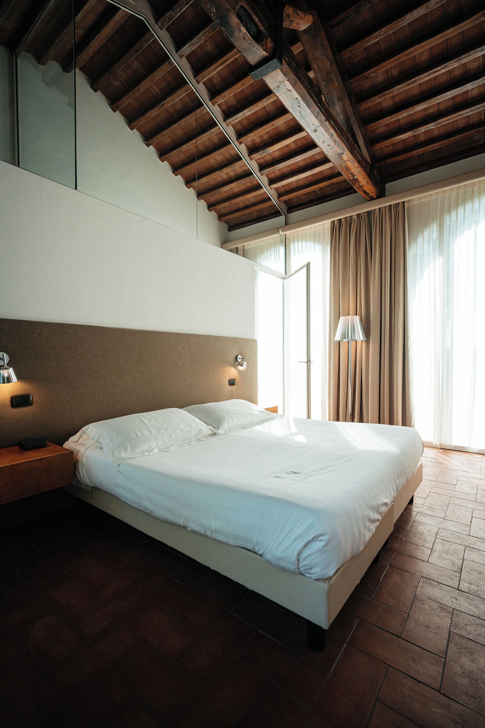A bedroom with a large bed, wooden ceiling beams, a nightstand, a lamp, and a window with curtains.