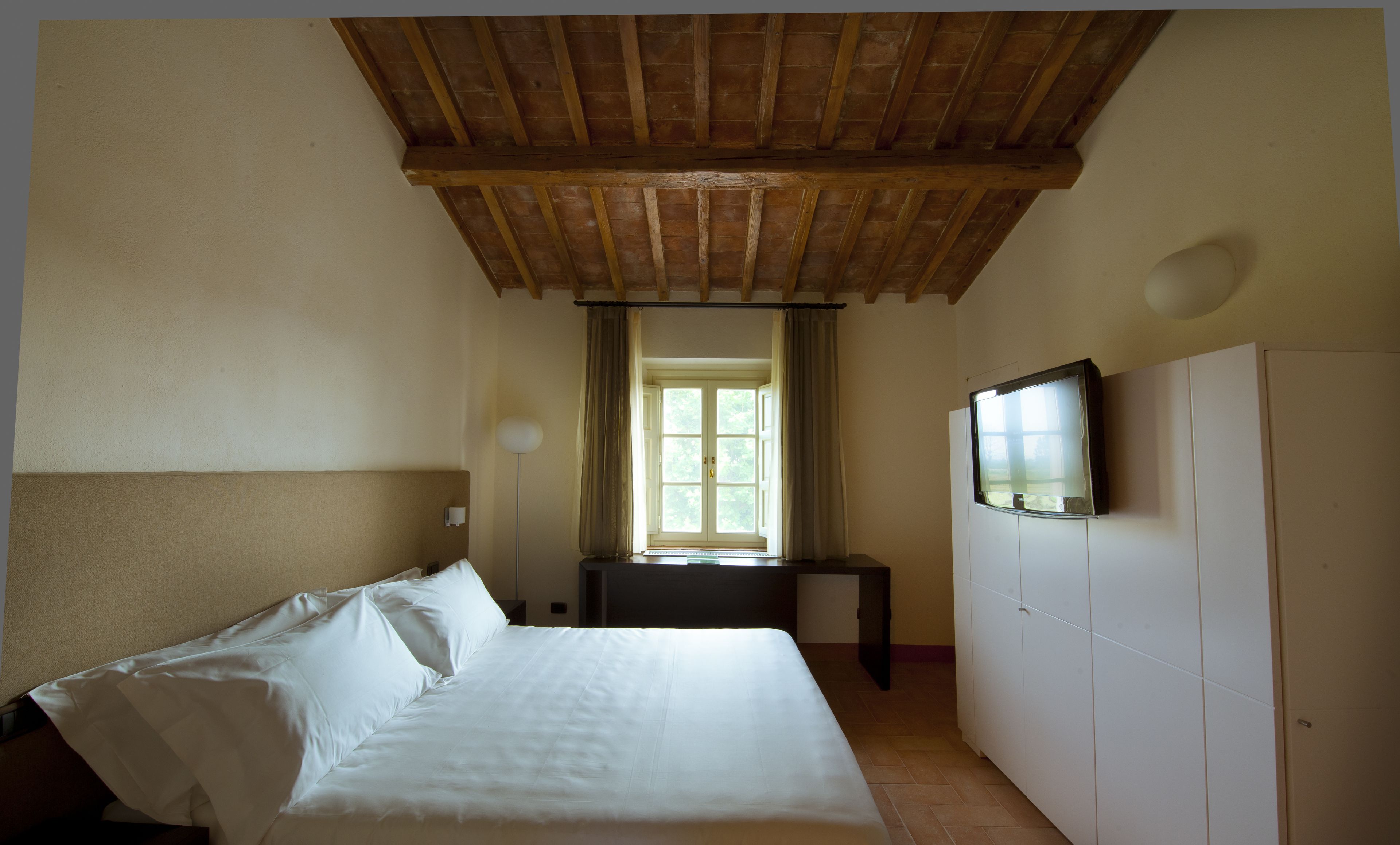 A hotel room with a wooden ceiling, a bed with white linens, a television on the wall, and a window with curtains.
