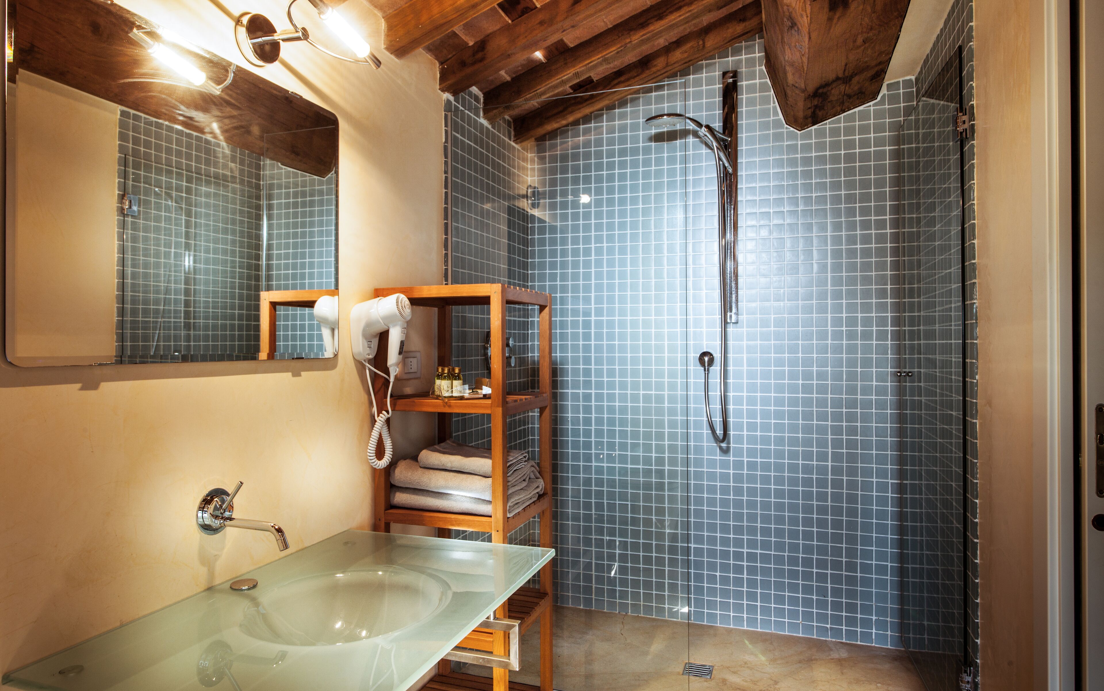 A bathroom with a glass sink, a wooden shelf with towels, a hairdryer, and a shower with blue tiles.