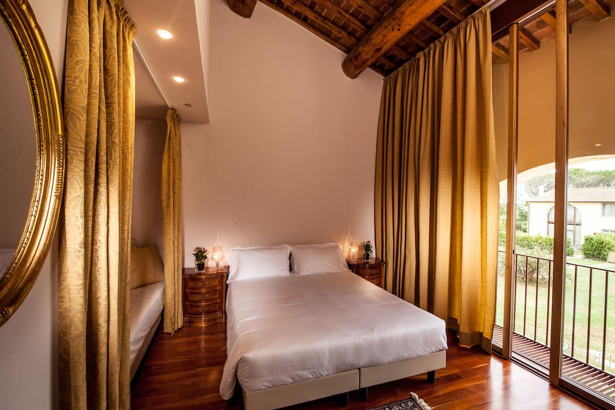 A bedroom with a large bed, wooden flooring, a ceiling with exposed beams, and a view of the outdoors through large windows.