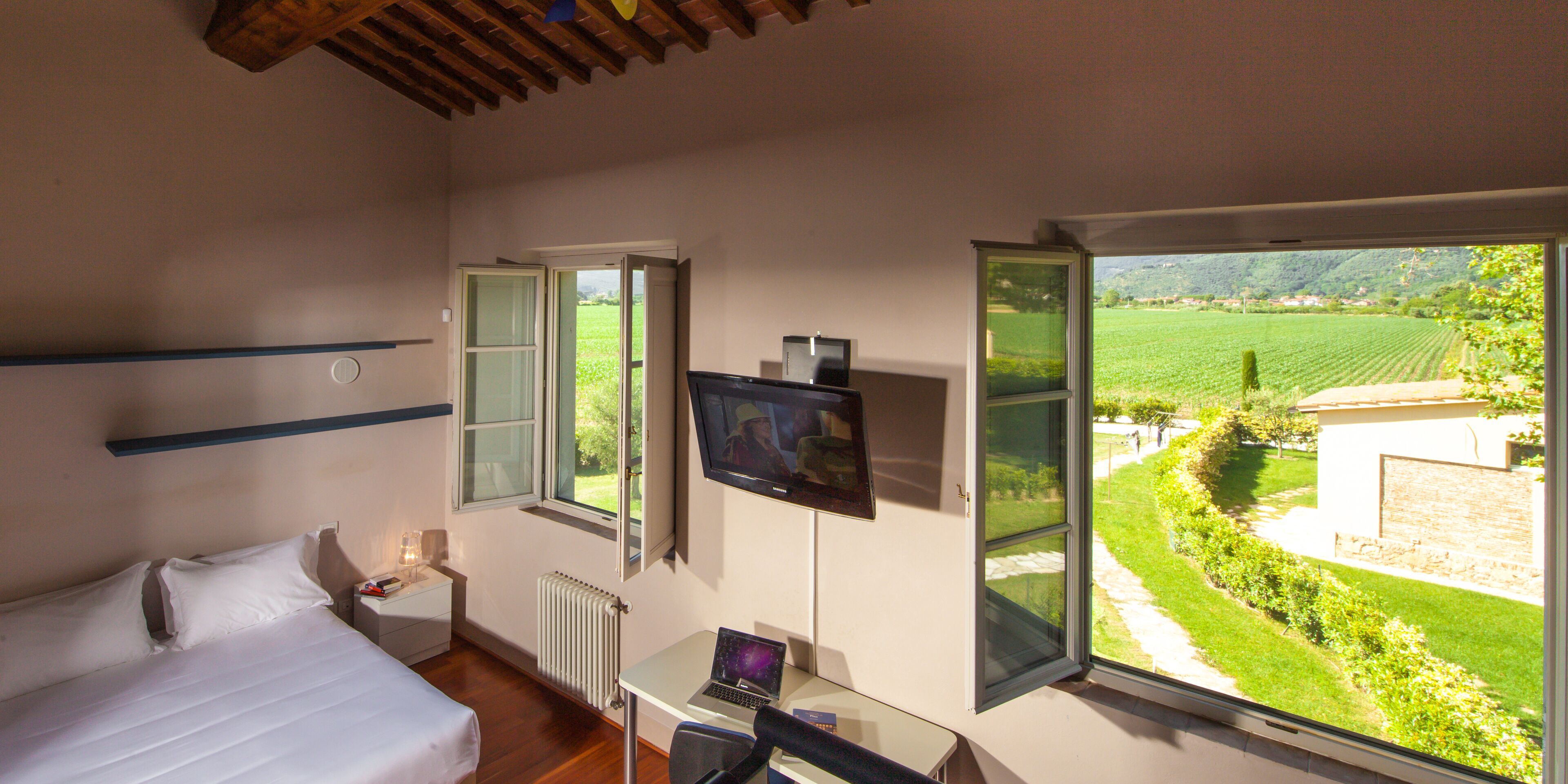 Een slaapkamer met een bed, een bureau met een laptop, een televisie en een raam met uitzicht op een groen landschap.