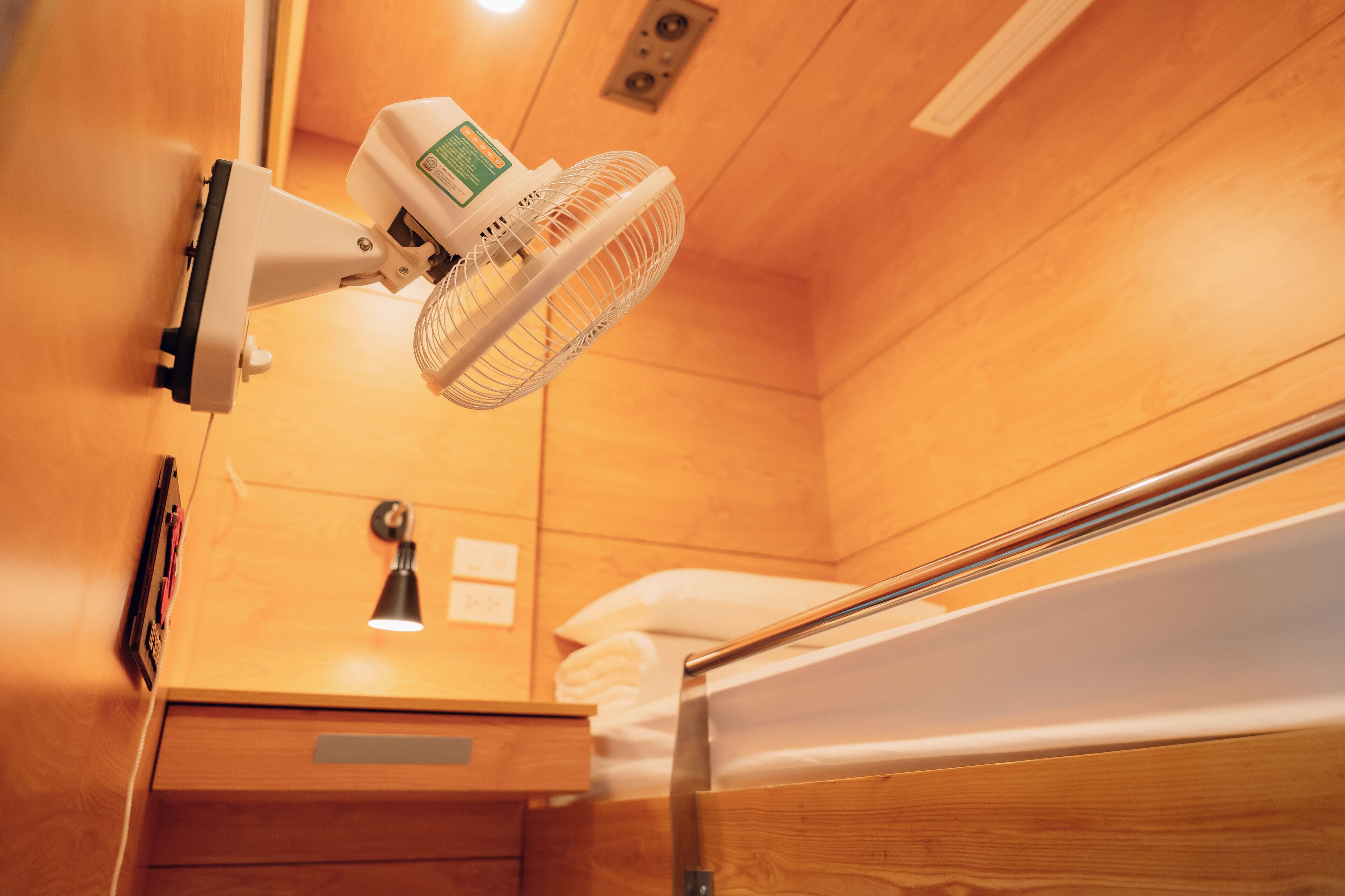 A wooden bunk bed with a bedside table and lamp, a wall-mounted fan, and a wooden wall panel.