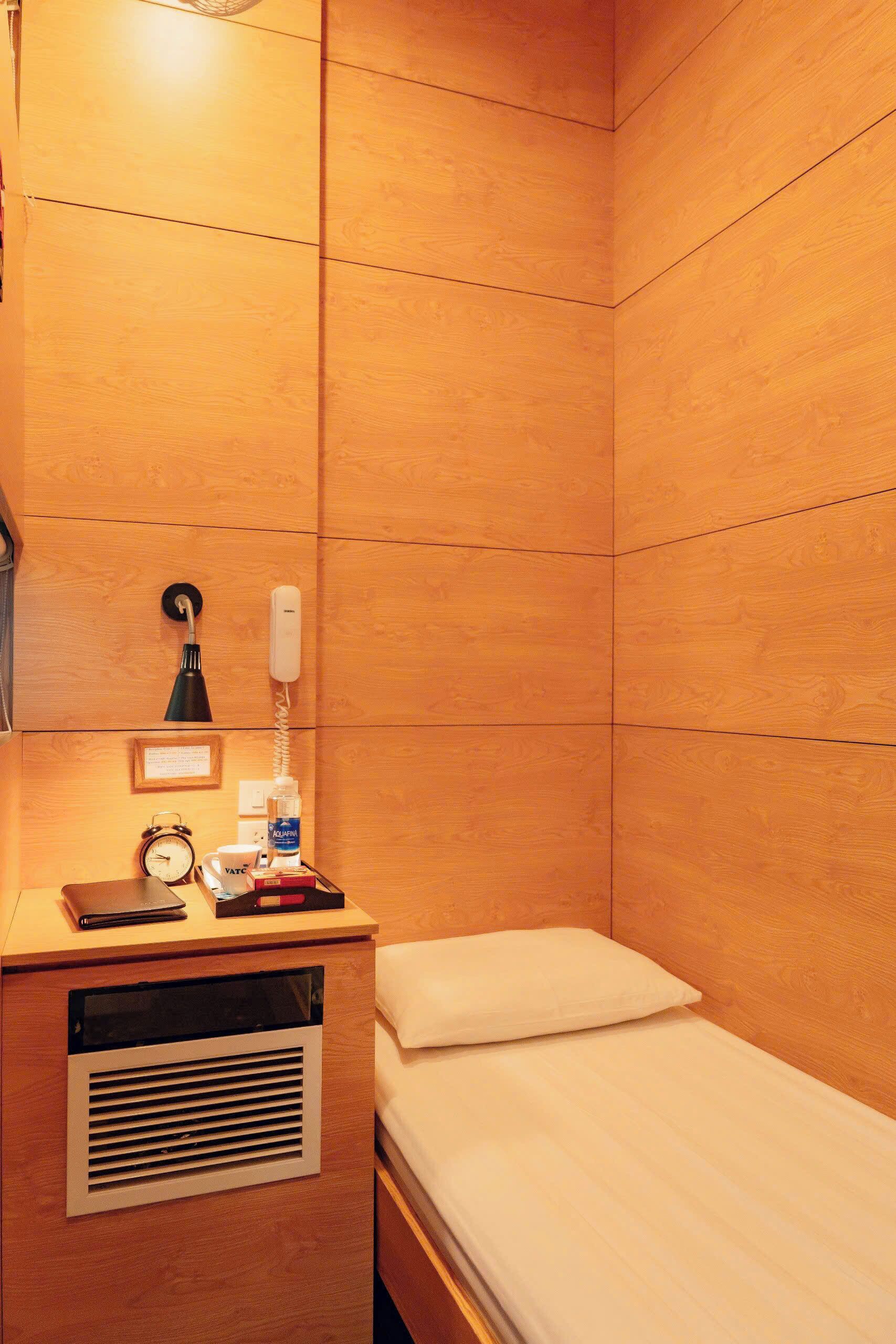 Une petite chambre avec un lit simple, des murs lambrissés, une table de chevet avec une lampe, un téléphone, une horloge et une bouteille d’eau.