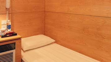 A small, single-bed room with a wooden wall, a bedside table with a lamp, a fan, and a phone.