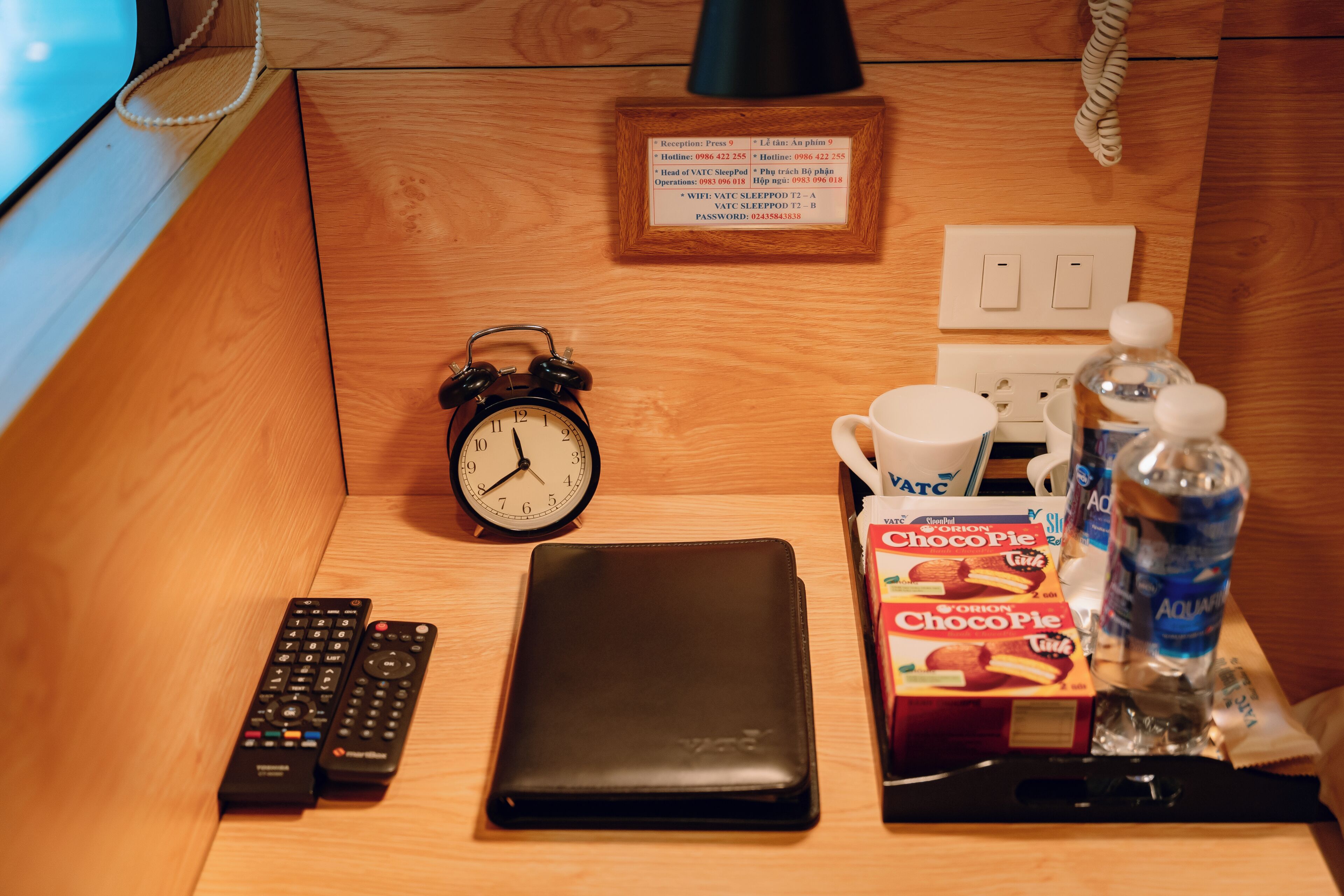 A wooden desk with a black alarm clock, two remote controls, a black wallet, a box of ChocoPie, a water bottle, and a cup.