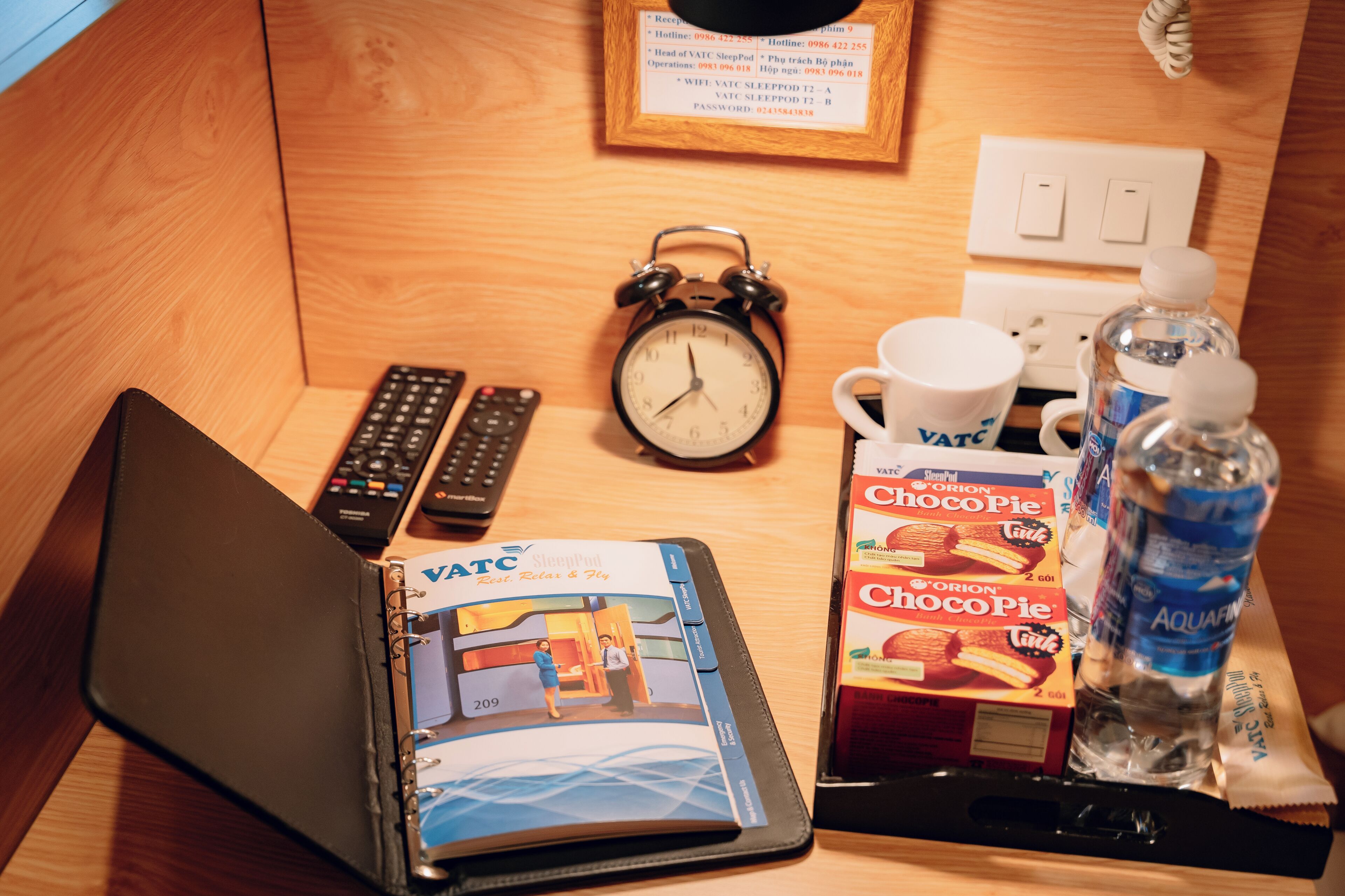 Ein Holztisch mit einem Laptop, einer VATC SleepPod, einer Fernbedienung, einer Kaffeetasse, einer Packung Chocopie, einer Wasserflasche und einem gerahmten Schild.