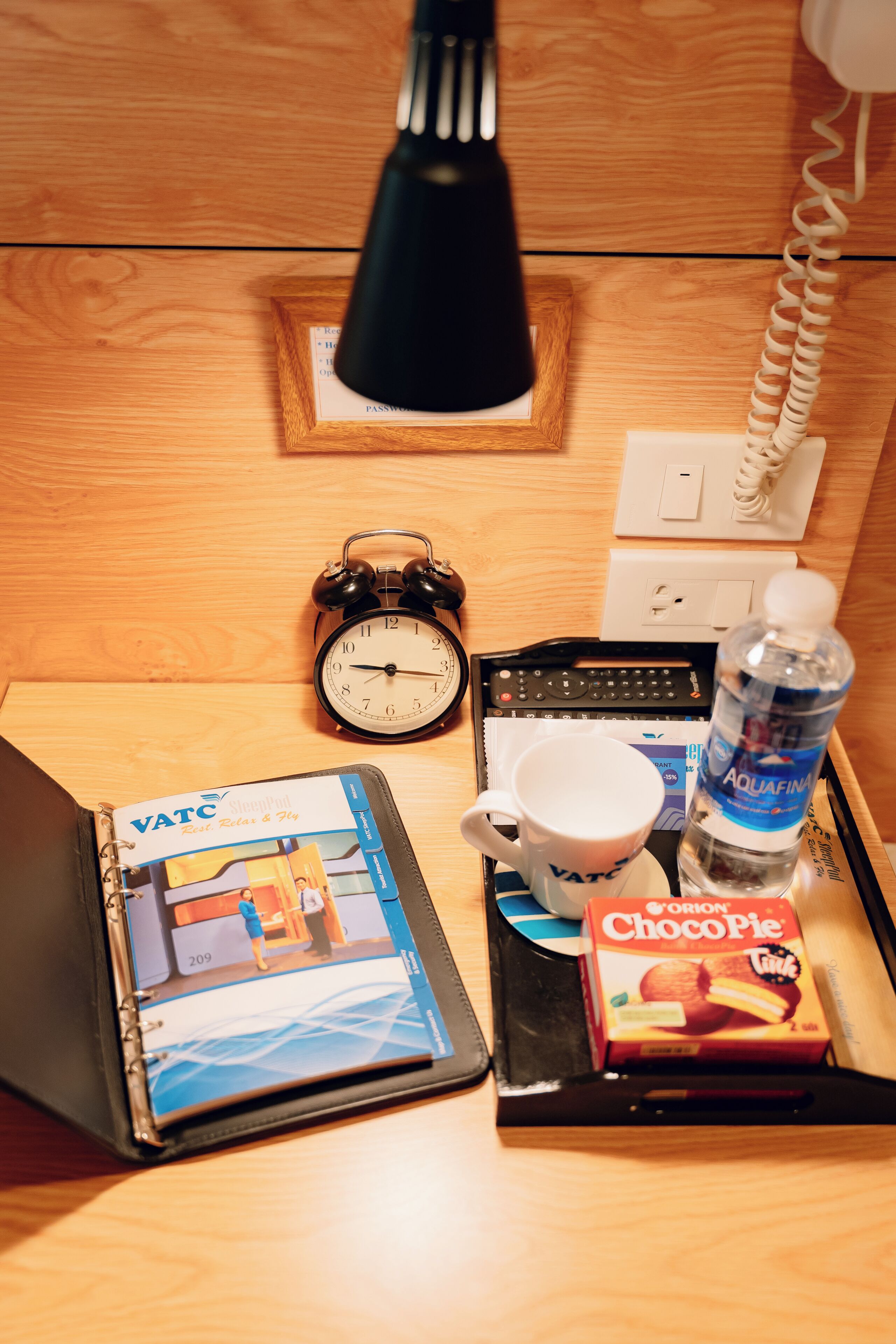 Ein Nachttisch aus Holz mit einer schwarzen Lampe, einem weißen Telefon, einem schwarzen Wecker, einem Laptop, einer Packung Orion ChocoPie, einer Wasserflasche und einer Tasse.