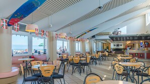 A spacious indoor dining area with multiple tables and chairs, large windows offering a view of the sea, and a bar area visible in the background.