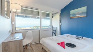A hotel room with a bed, a desk, a chair, a window with a view of the sea, and a painting on the wall.