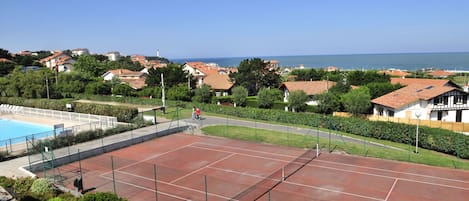 Una cancha de tenis con piscina, rodeada de edificios residenciales y una vista despejada del mar.