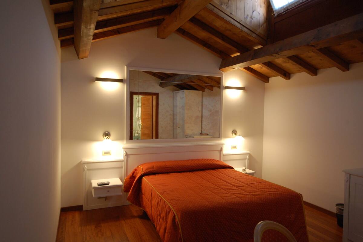 A bedroom with a wooden ceiling, a bed with an orange bedspread, two bedside tables with lamps, and a large mirror above the bed.
