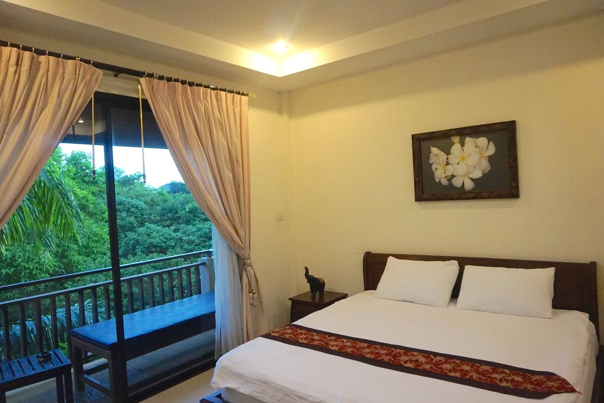 Quarto de hotel com cama, mesa de cabeceira, um banco e vista para o verde pela janela.