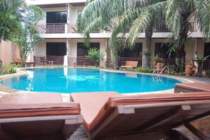 A swimming pool with lounge chairs, surrounded by palm trees and a multi-story building with balconies.