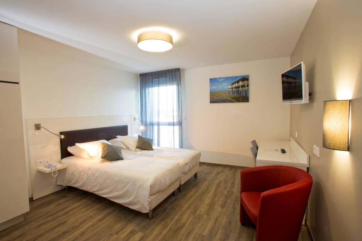 Ein Hotelzimmer mit zwei Betten, einem roten Sessel, einem Schreibtisch und einem Fernseher.