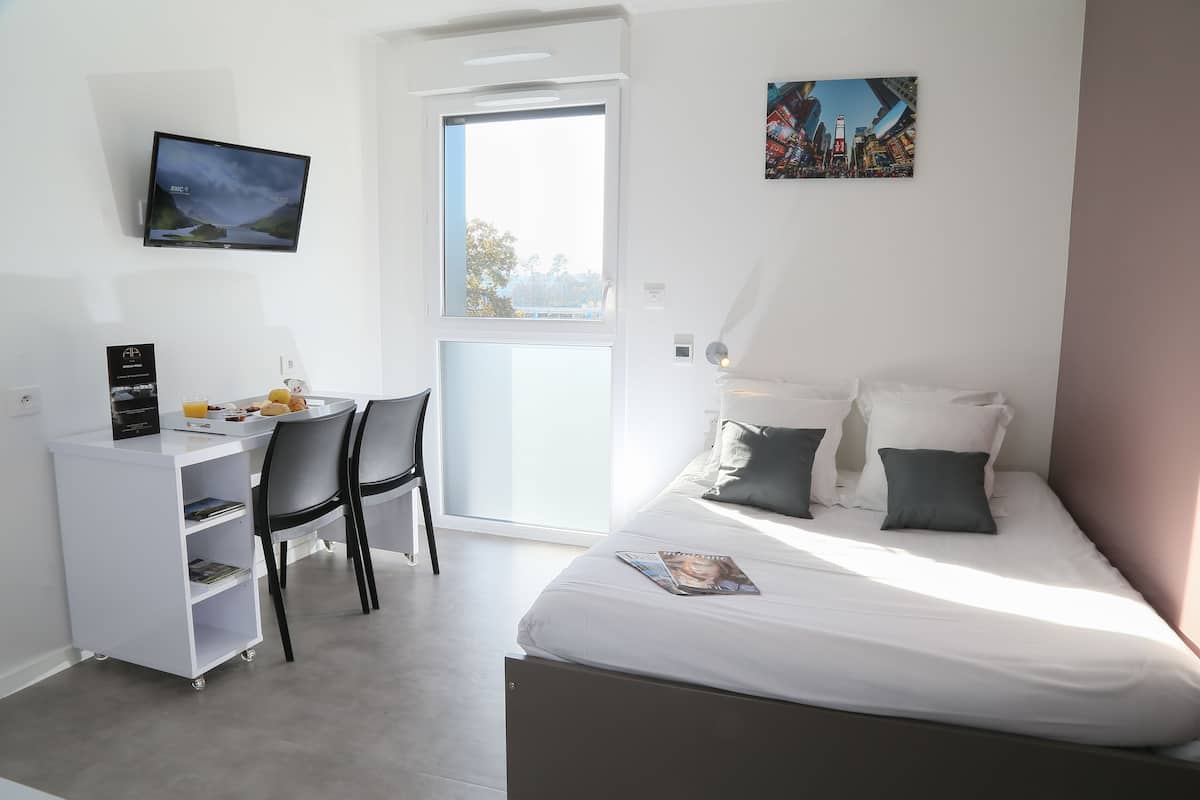 Ein modernes Schlafzimmer mit einem Bett, einem Schreibtisch mit Computer, einem Stuhl und einem Fenster.