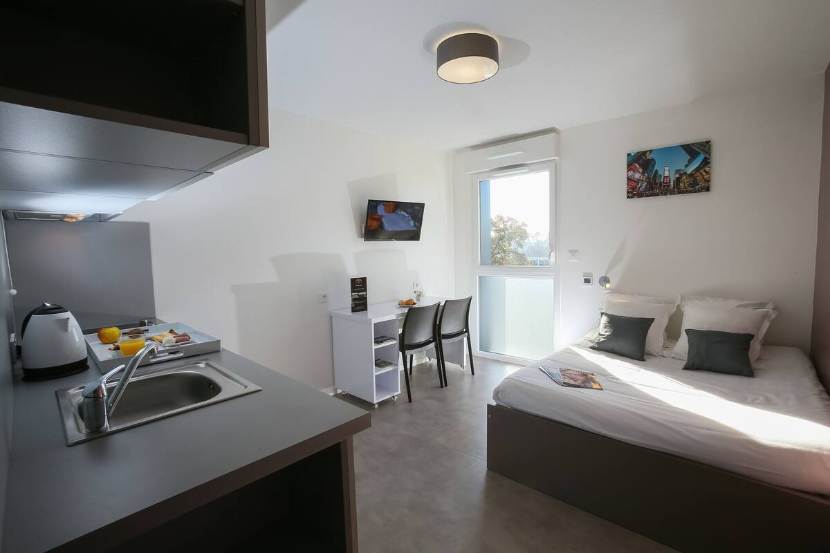 Ein modernes Hotelzimmer mit Küchenzeile, einem Bett mit Kissen, einem Schreibtisch mit Stuhl und einem an der Wand montierten Fernseher.