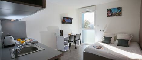 Habitación de hotel moderna con kitchenette, cama con ropa de cama oscura, escritorio con silla, televisor y una pintura de paisaje urbano.
