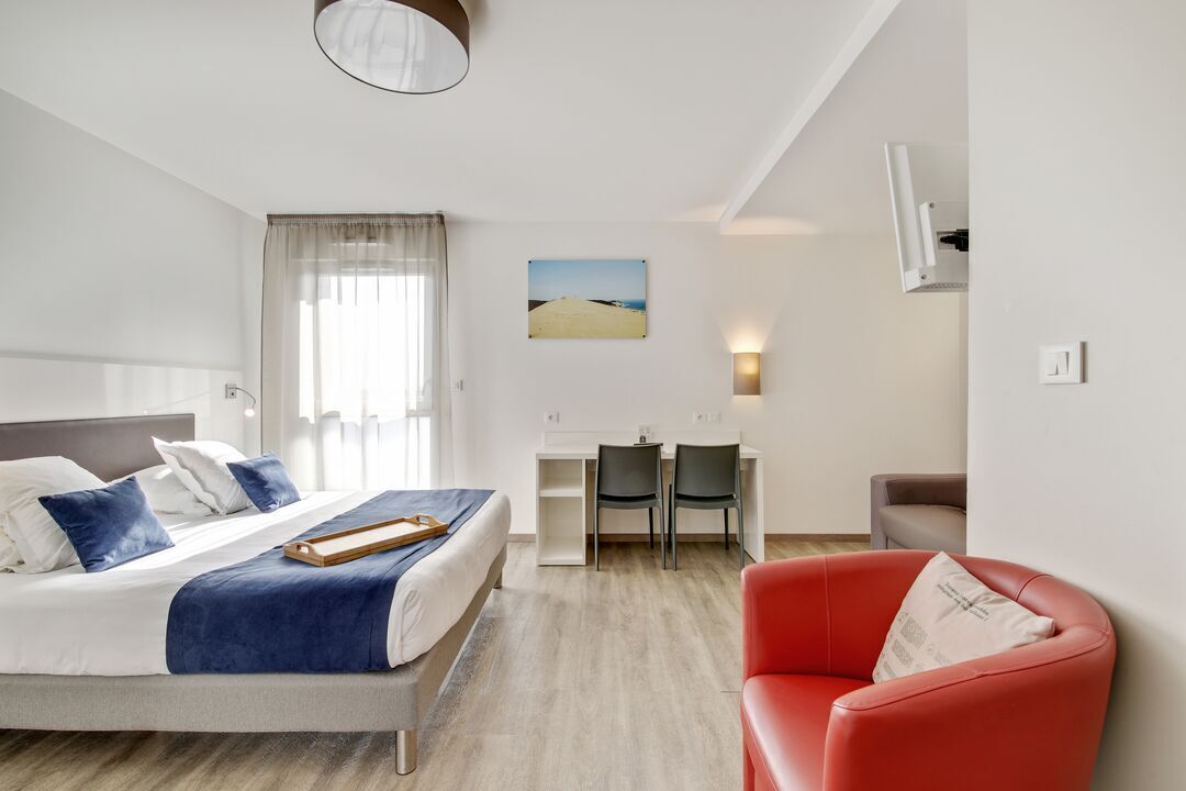 A hotel room with a bed, a desk, two chairs, a red armchair, and a painting on the wall.