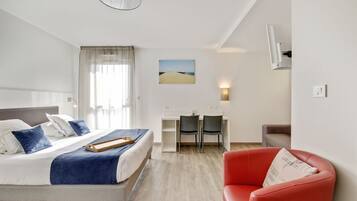 A hotel room with a bed, a desk, two chairs, a red armchair, and a painting on the wall.