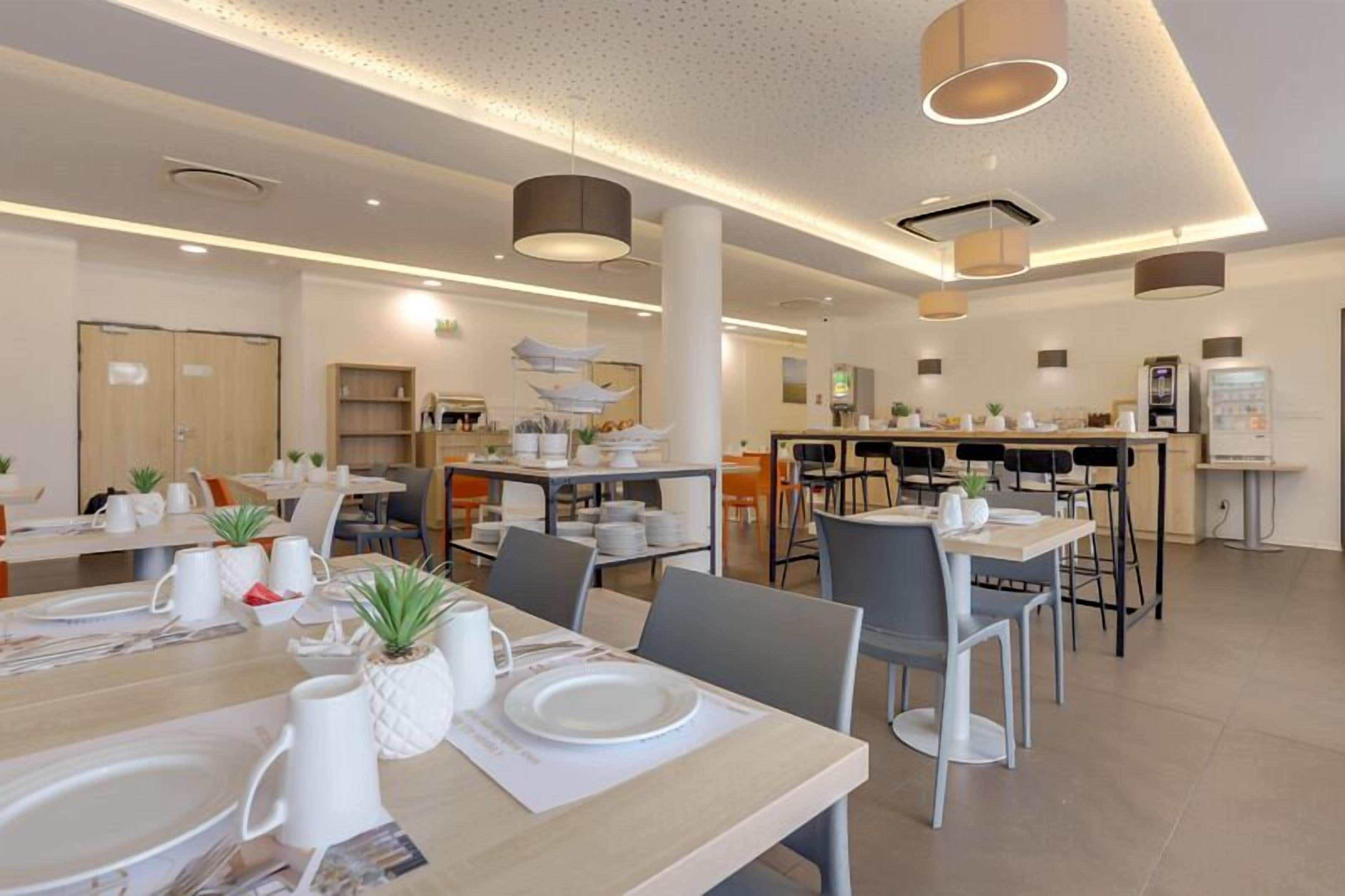 A modern hotel dining area with multiple tables and chairs, a kitchen in the background, and a clean, well-lit environment.