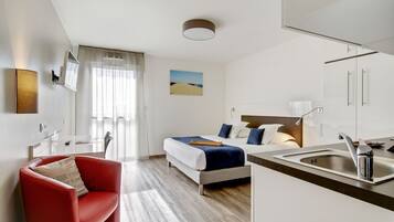 A modern hotel room with a bed, a red armchair, a kitchen area with a sink, and a large window with curtains.