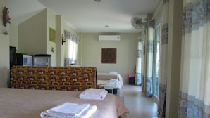 Ein Hotelzimmer mit einem großen Bett, einem Einzelbett, einer Couch und einem Fernseher. Es gibt ein Fenster mit Vorhängen, eine Klimaanlage und einen Ventilator.