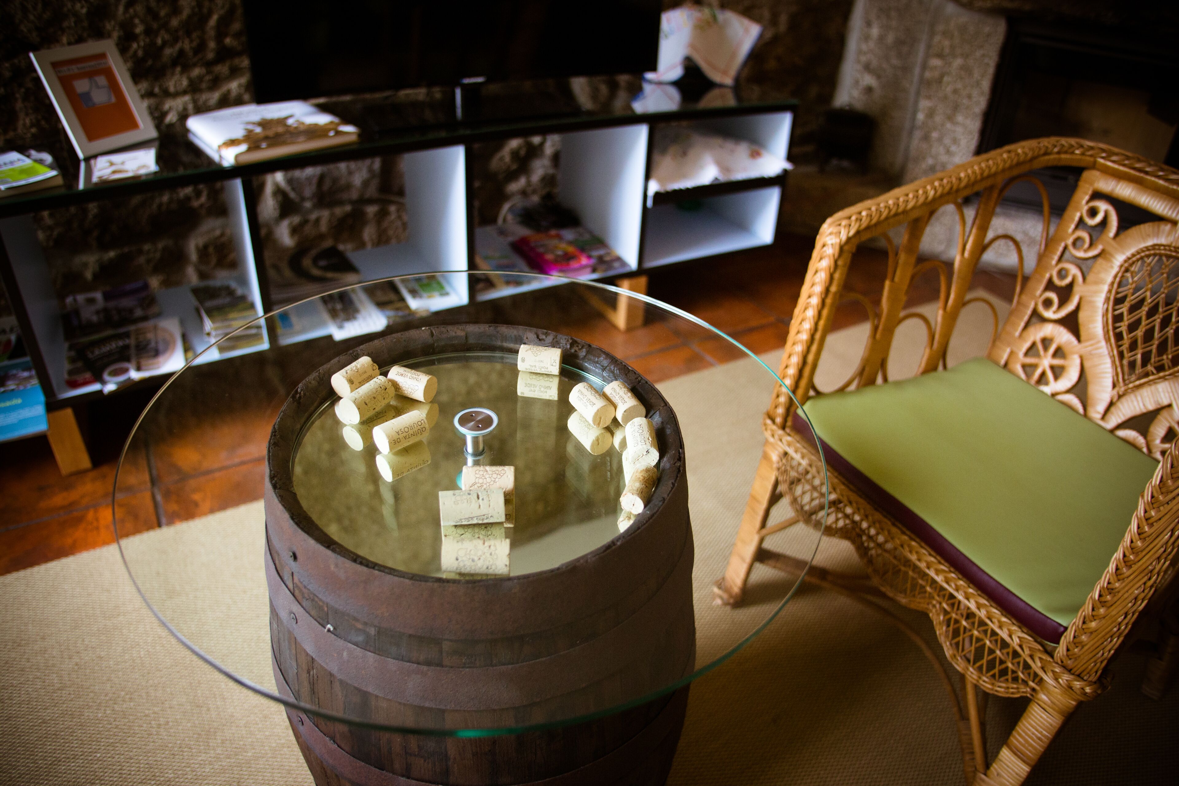 A round wooden barrel with a glass top, surrounded by corks. A wicker chair with a green cushion is to the right.