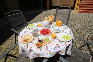 A round table set outdoors with a tablecloth, plates, cups, a teapot, a jug of orange juice, a bowl of cherries, and a wooden board with sliced apples and cheese.