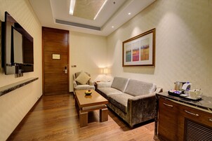 A hotel room with a wooden floor, a sofa, a small table with a fruit bowl, a coffee maker, and a framed picture on the wall.
