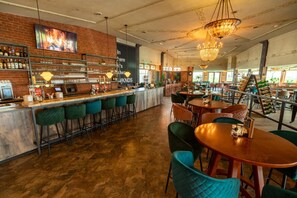 Una zona de bar con pared de ladrillo, estantes con botellas y una barra. Hay varias sillas y mesas, y un candelabro cuelga del techo.
