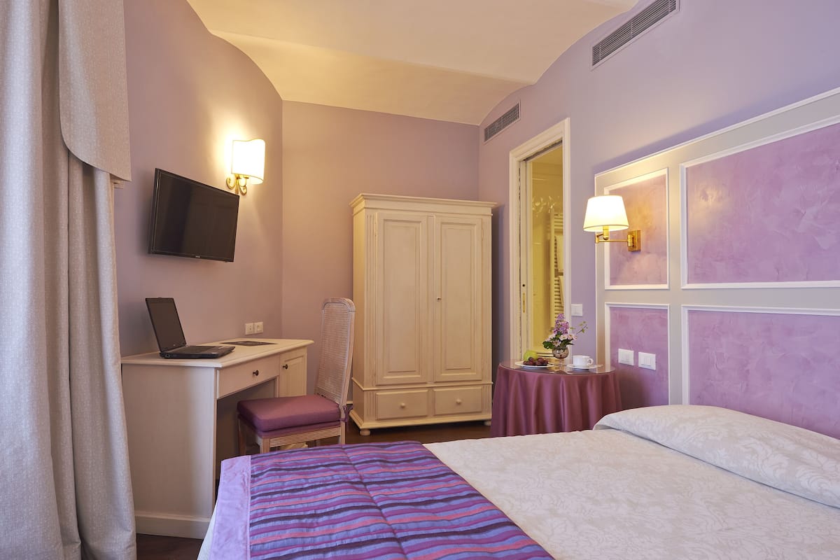 A hotel room with a bed, a desk with a laptop, a wardrobe, a small table with a vase, and a wall-mounted TV.