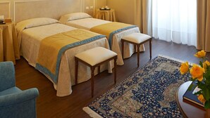 Una habitación de hotel con dos camas, un sillón azul, un banco de madera, un tapete estampado y un techo decorativo.