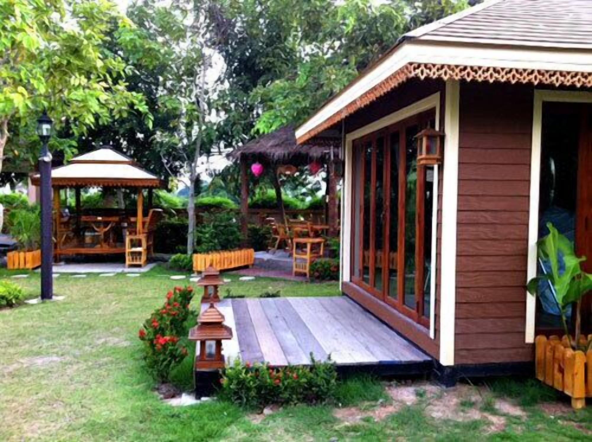 A wooden structure with a porch, surrounded by a garden with a gazebo and a thatched roof.