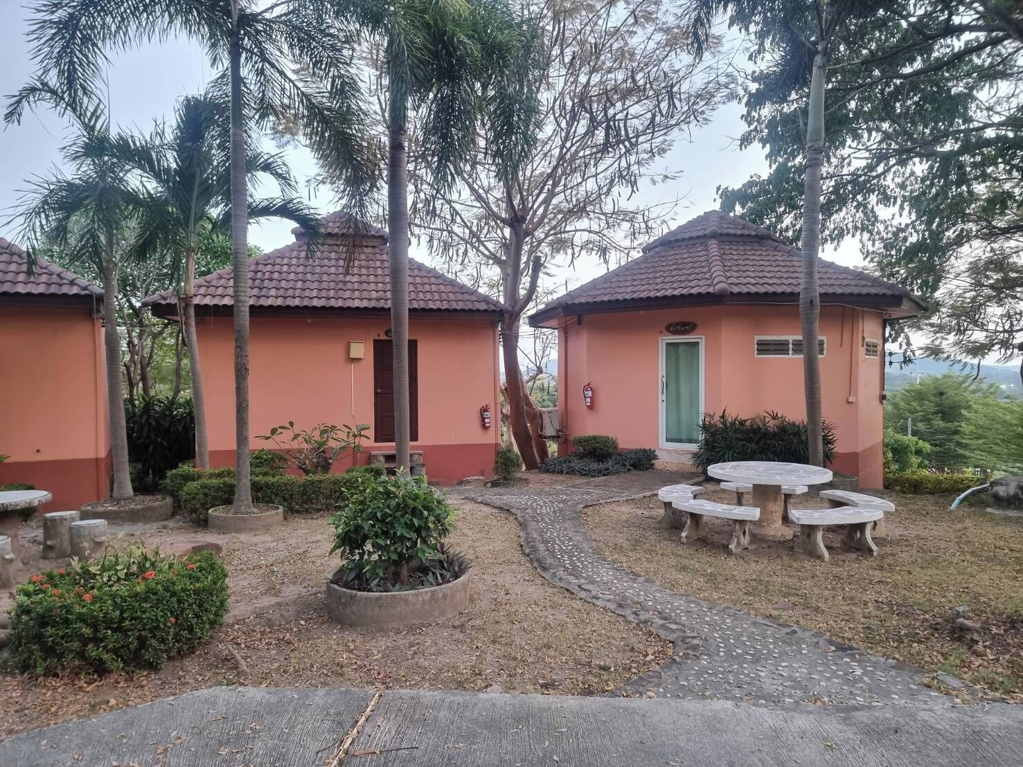 Two small, single-story buildings with red-tiled roofs, surrounded by palm trees and a gravel path leading to a circular seating area.