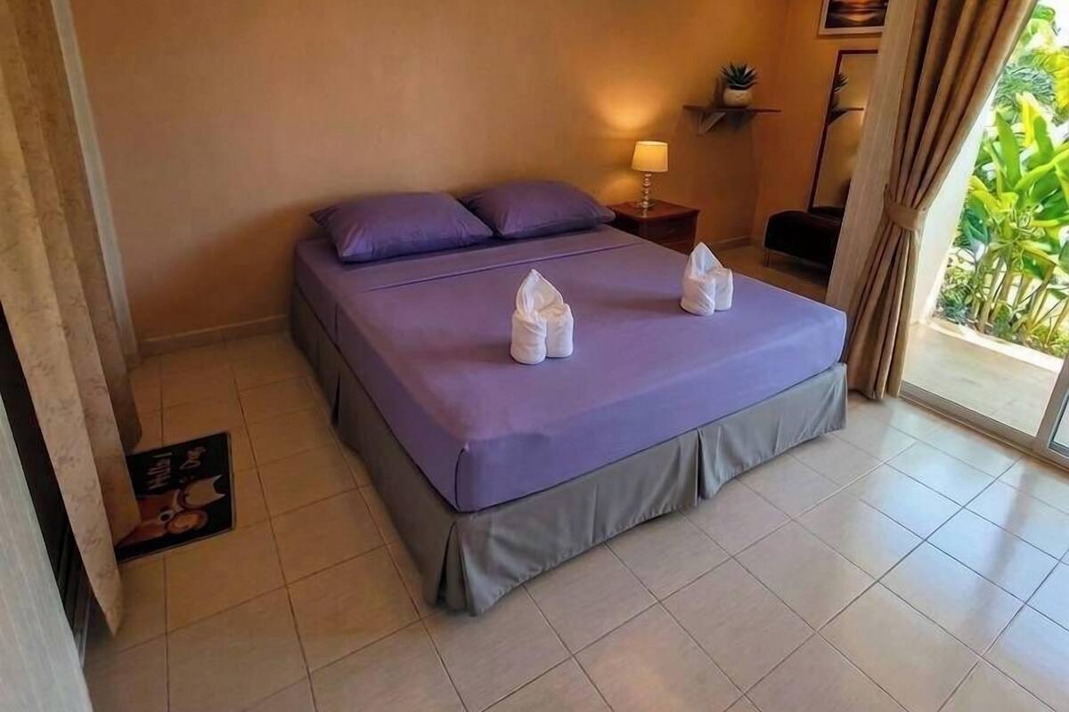 A hotel room with a bed, a bedside table with a lamp, a small shelf with plants, and a view of greenery through the window.