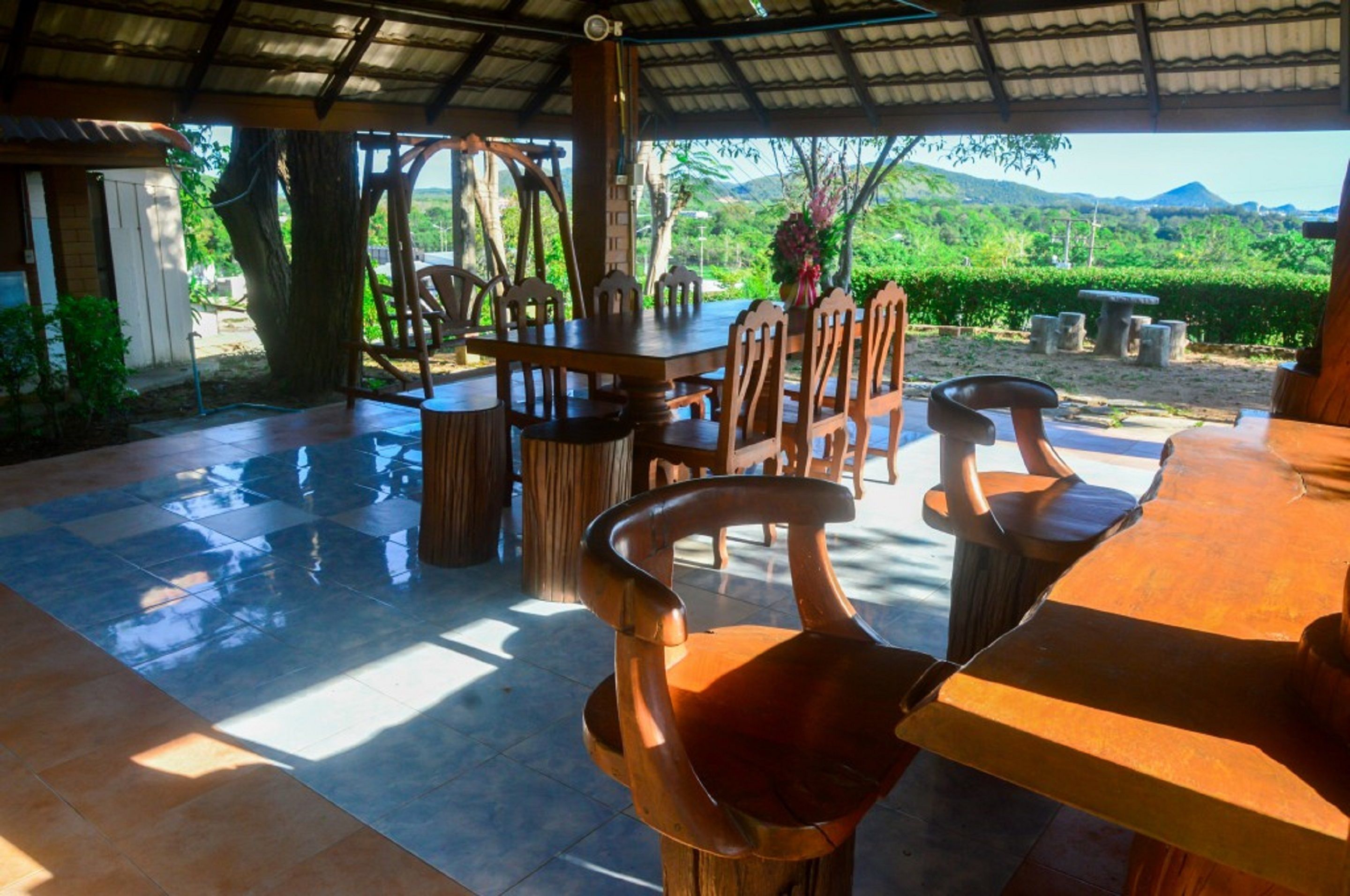 An outdoor dining area with a covered roof, wooden tables and chairs, and a view of lush greenery and mountains in the background.