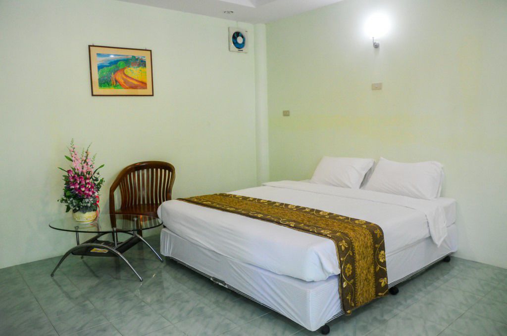 A single bed with a white mattress and a brown blanket, a wooden chair, a small glass table with a vase of flowers, and a wall-mounted fan.