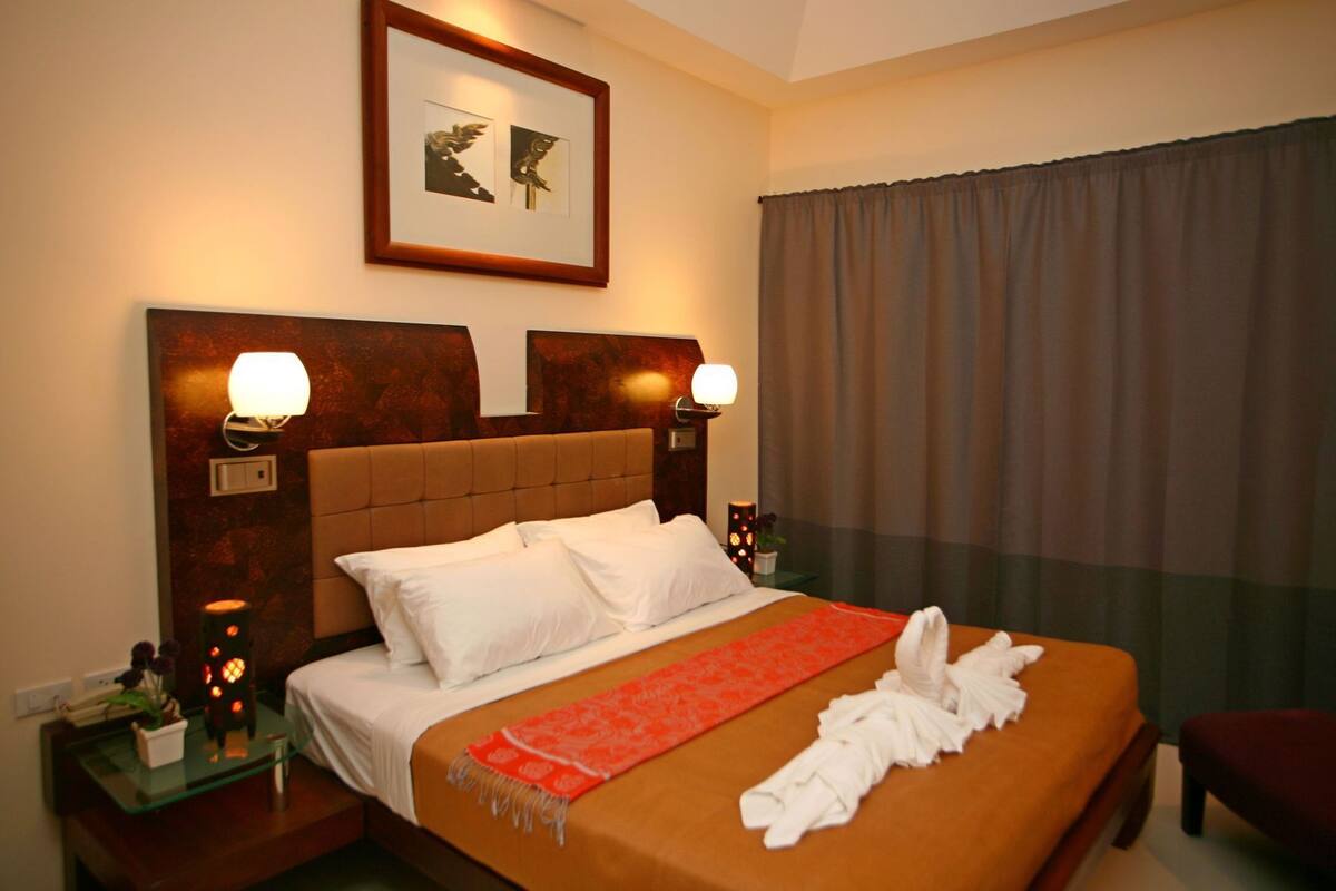 Ein Hotelzimmer mit einem großen Bett, zwei Nachttischlampen, einem gerahmten Bild über dem Bett, einem Fenster mit Vorhängen und einem kleinen Tisch mit einer Pflanze.