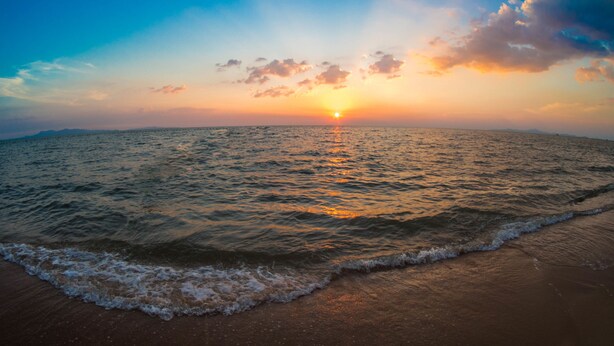 Una playa con olas que acarician suavemente la orilla, cielo despejado y el sol poniéndose en la distancia.