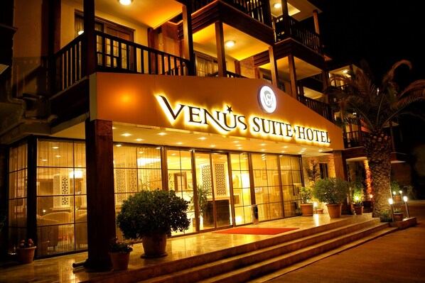Exterior del Hotel Venus Suite, con una entrada bien iluminada, ventanas amplias y plantas en macetas.