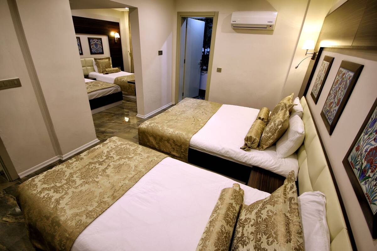 Quarto de hotel com duas camas, cada uma com colcha e almofadas estampadas. Há uma porta que dá para um banheiro, e obras de arte decoram as paredes.