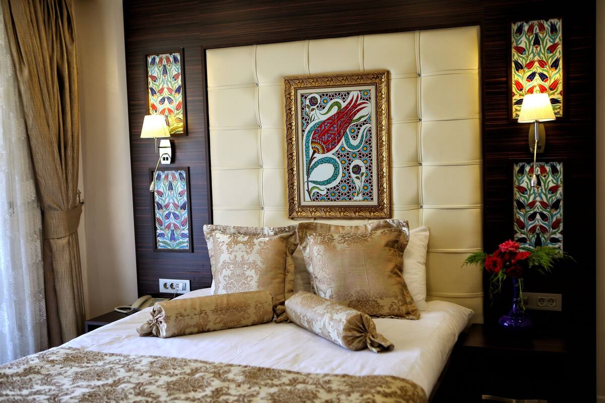 Quarto de hotel com uma cama grande, almofadas decorativas, cabeceira com uma obra de arte emoldurada, abajur e um vaso com flores.