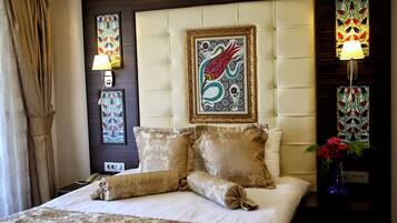 Una habitación de hotel con una cama grande, almohadillas decorativas, un cabecero con una obra de arte enmarcada, una lámpara de noche y un jarrón con flores.