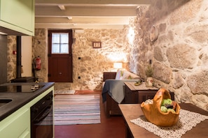 A cozy room with a stone wall, a wooden table, a sofa, a kitchen area with a stove, and a window with a door. - Traços D'Outrora (Vale de Cambra)