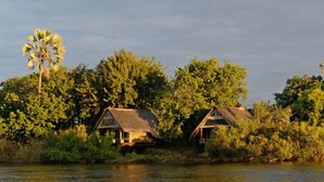 Deux charmantes chaumières au toit de chaume, situées au bord d’une rivière, entourées d’une végétation luxuriante et d’un palmier.