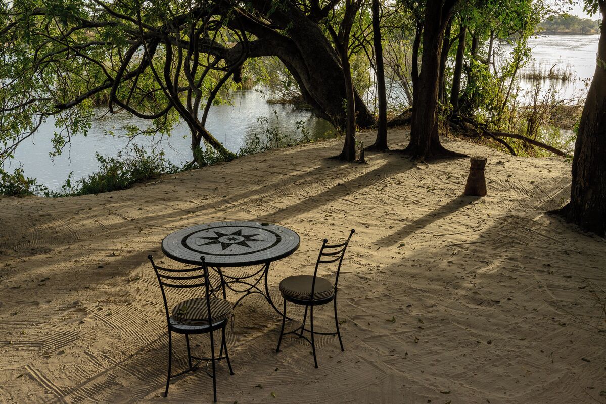 A round table with two chairs, a sandy area, and trees near a body of water.